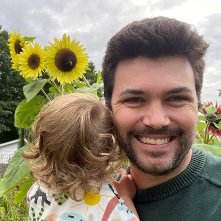 Shane Scranton smiling in front of sunflowers, holding a toddler